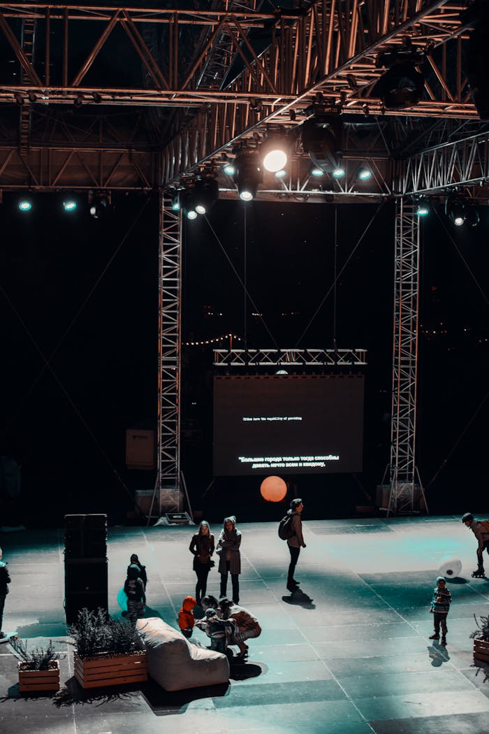 A vibrant nighttime stage setup with a crowd, lights, and a projected screen creating a lively atmosphere.